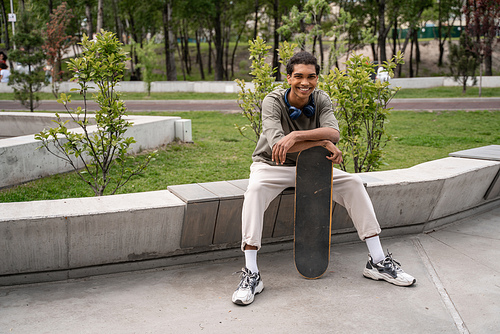 cheerful african american man smiling at camera while sitting on border bench with skateboard