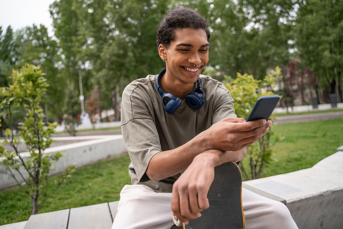 joyful african american skater sitting on border bench and messaging on smartphone