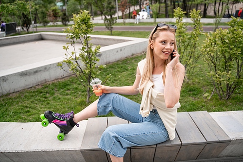 smiling roller skater with cocktail sitting on border bench and talking on mobile phone