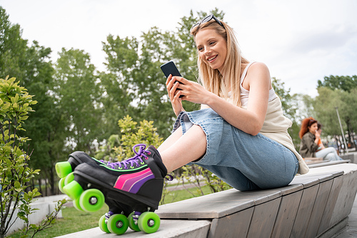 happy blonde woman in rollers skates sitting on border bench and messaging on cellphone