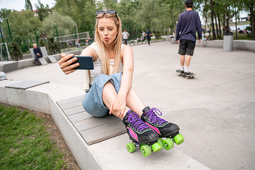 young woman in rollers skates taking selfie on cellphone in skate park