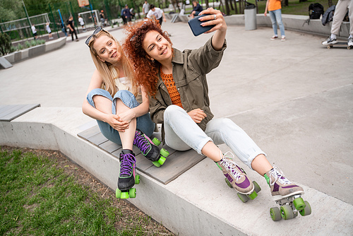 cheerful friends in rollers skates taking selfie on border bench in skate park