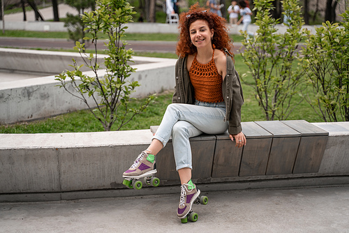 happy and curly roller skater smiling at camera on bench in skate park