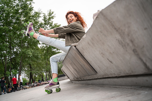 low angle view of cheerful woman tying laces on roller skate while sitting on border bench