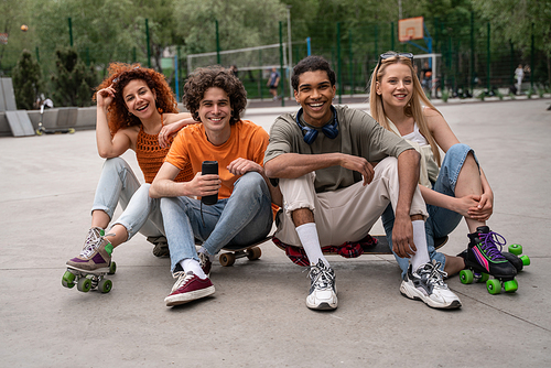 happy multiethnic skaters sitting on asphalt in skate park with portable music speaker