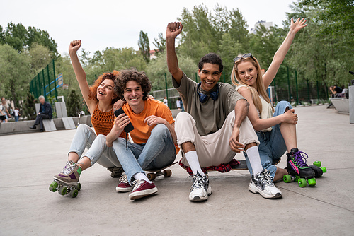 excited multicultural skaters showing win gesture while sitting on road in skate park