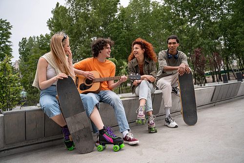 curly man playing guitar to happy multiethnic friends in skate park