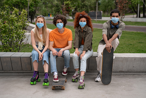 multiethnic friends in medical masks sitting on border bench in skate park