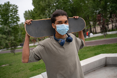 young african american man in medical mask holding skateboard behind neck and 