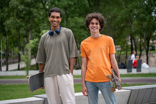 joyful interracial friends with skateboards smiling at camera outdoors