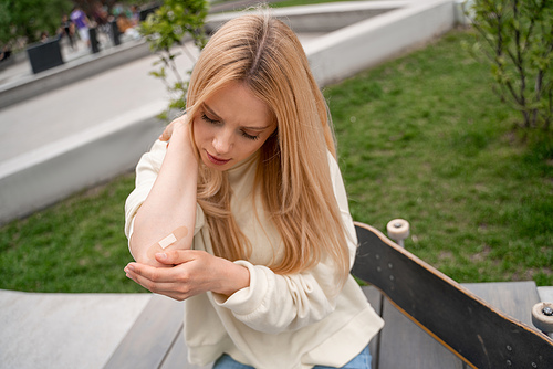 blonde woman applying adhesive plaster on injured elbow near skateboard on bench