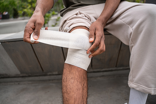 cropped view of african american man sitting on border bench and bandaging injured knee