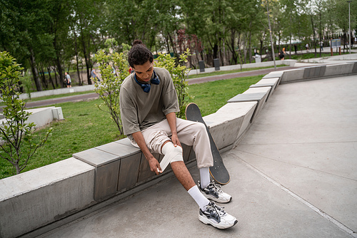 young african american skater bandaging injured knee while sitting on border bench