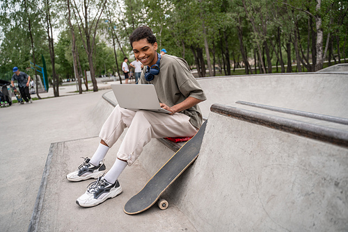 smiling african american student sitting on border near skateboard and using laptop