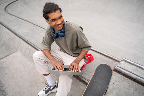 high angle view of cheerful african american student using laptop on border near skateboard