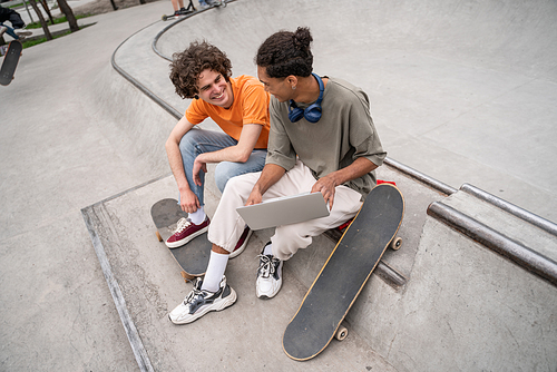 happy interracial skaters talking while sitting on skate ramp with laptop