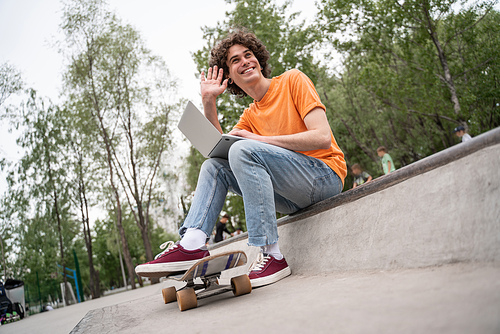 cheerful skateboarder with laptop waving hand while sitting outdoors