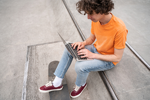 young curly skateboarder typing on laptop in skate park