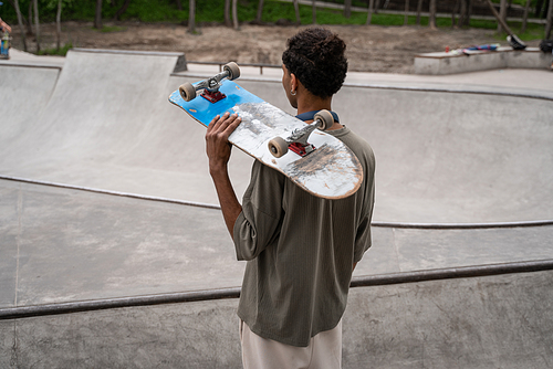 african american man with skateboard on shoulder looking at skate ramp