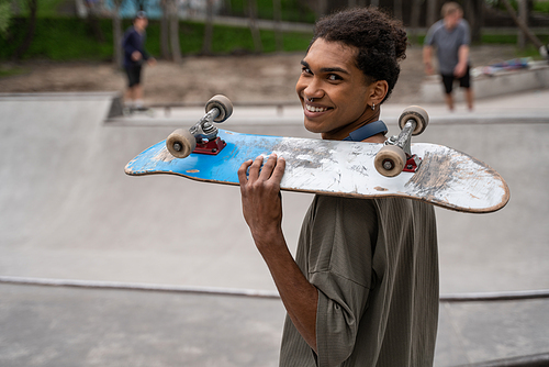 young african american man smiling at camera while holding skateboard on shoulder