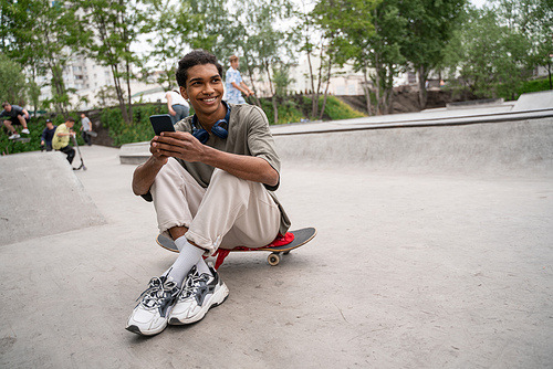 young and happy african american man using smartphone while sitting on skateboard