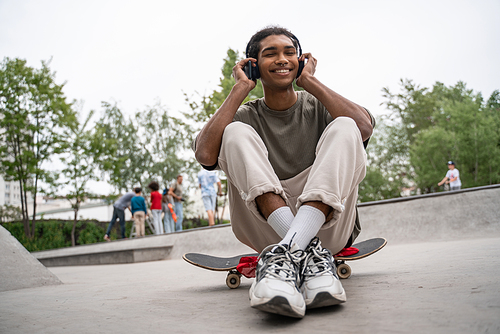 joyful african american man listening music in headphones while sitting on skateboard