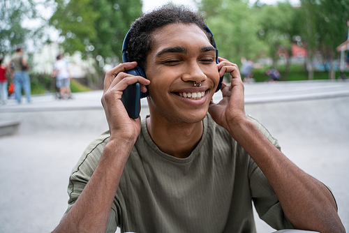 pleased african american man listening music in wireless headphones in city park