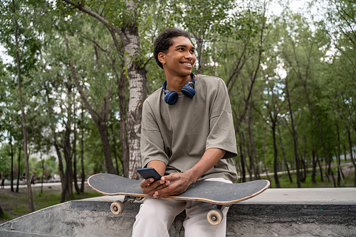 cheerful african american man with headphones, skateboard and mobile phone sitting on border outdoors