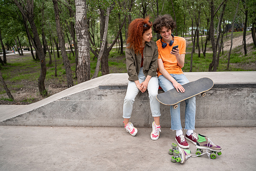 young skater using smartphone while sitting with friend on border in park