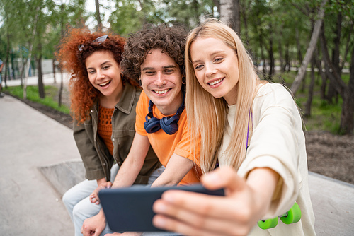 smiling blonde woman taking selfie with happy friends in park
