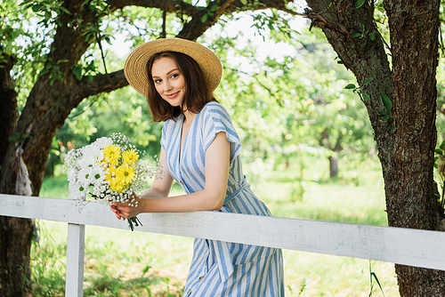 Woman in straw hat holding flowers and smiling at camera near fence