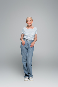 full length view of senior woman posing with thumbs in pockets on grey