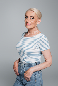 grey haired woman in striped t-shirt posing with thumbs in pockets of jeans isolated on grey