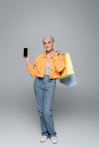positive elderly woman posing with shopping bags and mobile phone on grey