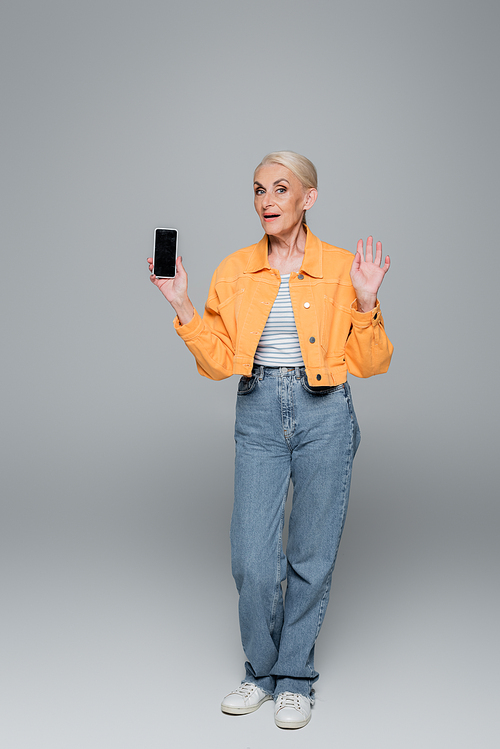 stylish senior woman waving hand and showing smartphone with blank screen on grey