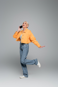 excited and stylish senior woman talking on smartphone while standing on one leg on grey