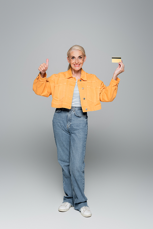 elderly woman in jeans and orange jacket showing credit card and like sign on grey