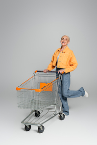 cheerful elderly woman in stylish clothes standing on one leg near shopping cart on grey