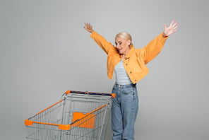 excited senior woman showing wow gesture while looking at shopping cart on grey