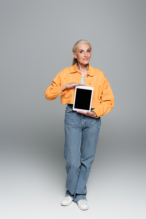 elderly woman in trendy clothes showing digital tablet with blank screen on grey