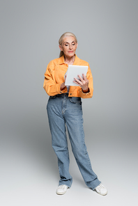 full length view of senior woman in jeans and jacket using digital tablet on grey