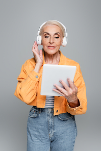 stylish senior woman looking at digital tablet while adjusting headphones isolated on grey