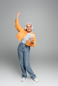 joyful senior woman in headphones holding digital tablet and showing win gesture on grey