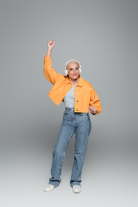 full length view of senior woman in jeans and jacket dancing in headphones on grey