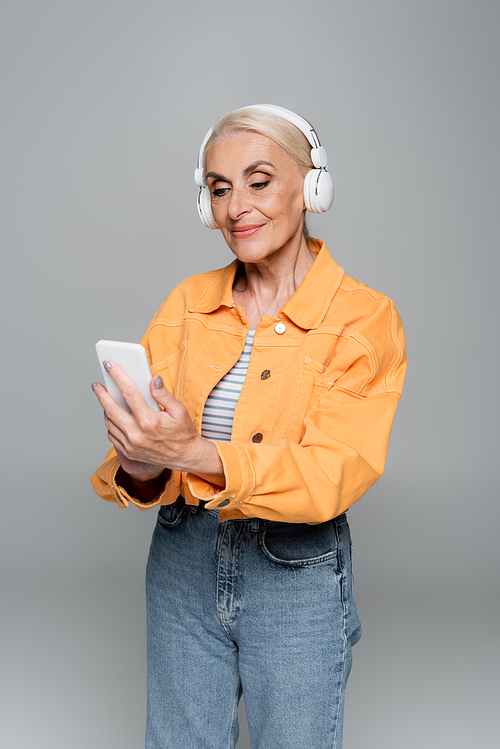 positive senior woman in headphones using mobile phone isolated on grey