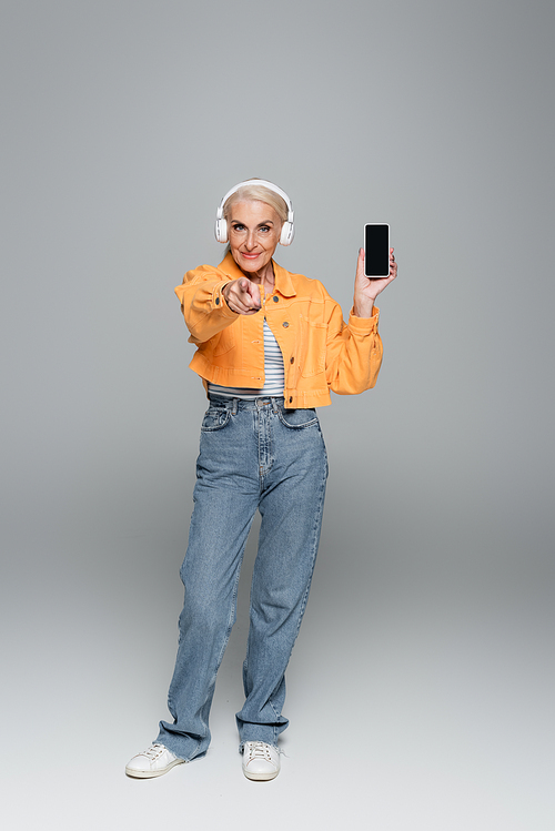 elderly woman in stylish clothes and headphones holding cellphone with blank screen on grey