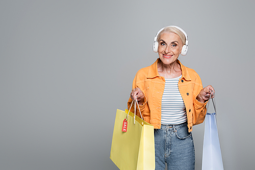 happy senior woman in headphones holding shopping bags isolated on grey