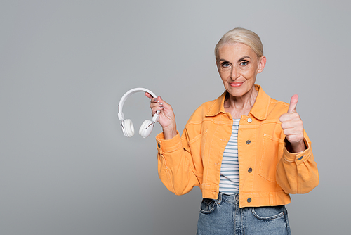 elderly woman in orange jacket showing thumb up and headphones isolated on grey