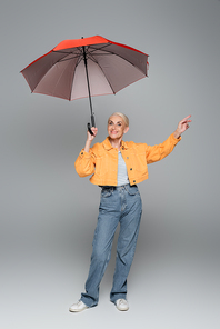 joyful woman in orange jacket and jeans posing under umbrella on grey