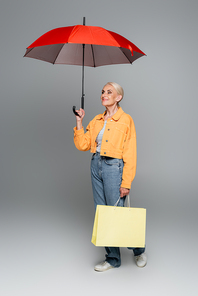 senior woman in jacket and jeans standing with shopping bag and smiling under umbrella on grey